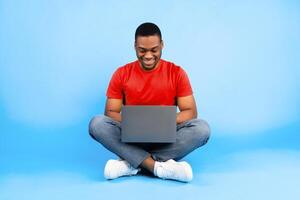 Happy Black Man Using Laptop Working Online Sitting Over Blue Background In Studio. Successful African American Male Freelancer Or Blogger Guy Browsing Internet On Computer photo