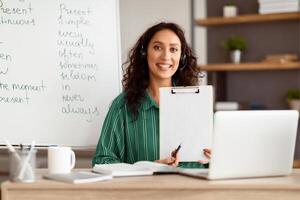 Smiling lady manager having videocall, looking at camera, giving virtual presentation or online lesson to students. Woman showing document contract to webcam, pointing to blank empty sheet of paper photo