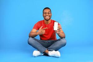 Smiling African Man Showing His Mobile Phone With Empty Screen Recommending Great App For Smartphone Sitting Over Blue Studio Background. People And Gadgets, New Application. Mockup photo