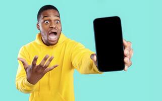 Emotional Black Guy Showing Big Smartphone With Empty Screen To Camera Advertising New Amazing Mobile Application Over Blue Studio Background. Shocking News, Surprising App For Your Phone photo