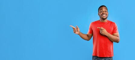 Look Aside. Black Male Pointing Fingers Showing Copy Space For Advertisement Text Standing Wearing Yellow T-Shirt On Blue Studio Background, Smiling To Camera. Panorama photo
