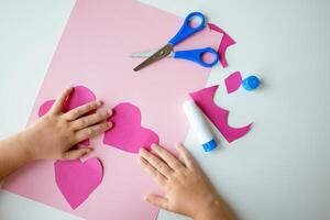 Childs hands making pink paper hearts with scissors and glue on white table photo