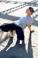 A young man practices a dance move on a sunny day, his long shadow stretching across the paved ground photo