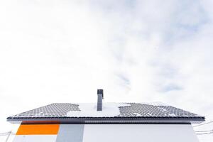 Chimney adorns snow-covered roof in city skyline amidst cumulus clouds, buildings, and sloping facades. photo