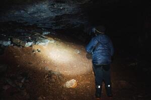 A solitary person stands inside a dark, eerie cave, grasping a flashlight that illuminates the surrounding area while shadows playfully dance and flicker on the rocky walls around them photo