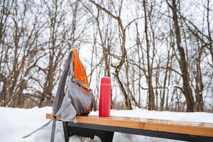 On a chilly winter day, a backpack and an insulated thermos are resting on a bench, surrounded by a blanket of soft, white snow, creating a picturesque and serene winter scene photo