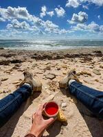 A person is leisurely sitting on the beach while holding a cup filled with a refreshing red liquid, enjoying the wonderful ambiance of summer by the seaside, with the sun shining brightly photo