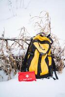 A bright yellow backpack is resting quietly in the soft, white snow, positioned right next to a striking red first aid kit, creating a vivid contrast in this snowy landscape photo