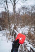 A person is warmly dressed in insulated clothing and is holding a bright red first aid kit while standing comfortably in soft, powdery white snow that blankets the ground all around them photo