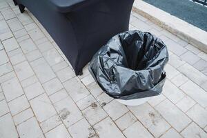 A black trash bag next to a sturdy wooden table highlights the contrast between waste and functionality in an outdoor setting, showcasing discarded items versus functional furniture photo