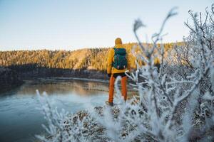 Back view of a man with a backpack standing on the edge of a cliff against the background of mountains, a guy is traveling alone, a man is standing on a mountain, looking forward at a pond. photo