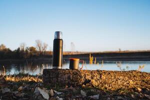 A classic thermos and a ceramic cup sit gracefully together on a smooth rock near a tranquil lake, surrounded by breathtaking scenery, beneath a clear blue sky and gentle, lapping waves photo