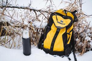 en el sereno invierno paisaje, un vibrante amarillo mochila descansa en el suave blanco nieve, acompañado por un robusto negro termo, creando un sorprendentes y hermosa contraste foto