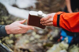 Man in orange jacket gesturing as he hands a flask to another man photo