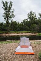 Imagine the tranquility of camping next to a calm riverside in nature, surrounded by greenery with a tent on a pebble shoreline, offering relaxation and a scenic view photo