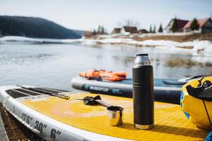 A thermos with a mug stands on a SUP against the background of a water landscape, a hot drink in cold weather, a tourist rafting on a SUP board. photo