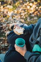 A person is comfortably sitting on the ground, enjoying a delicious sandwich while simultaneously sipping on a warm cup of coffee, taking in the surrounding scenery of nature photo
