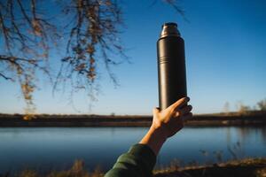 A person stands by a serene lake, holding a thermos in their hand, gazing over the calm water, surrounded by lush plants and trees under a beautiful sky filled with clouds photo