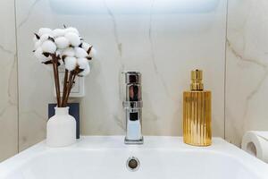 In a stylish bathroom, there is a lovely sink alongside a vibrant vase filled with beautiful cotton flowers and a welldesigned soap dispenser for added convenience and elegance photo
