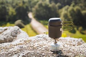 A compact camping stove set up on a rugged rocky surface, providing warmth and cooking capabilities while overlooking a scenic trail photo