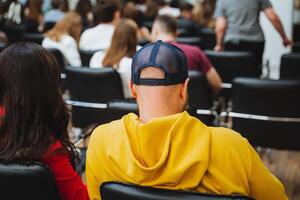 a man in a yellow hoodie is sitting in a chair in front of a crowd of people photo