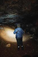 A person is standing alone in a dimly lit cave, holding a flashlight that illuminates the surroundings, casting electric blue hues against the darkness of the expansive landscape photo