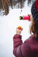 A woman is carefully pouring steaming coffee from a thermos into a cup while standing in the snow, surrounded by a winter wonderland of white, chilly flakes falling gently around her photo