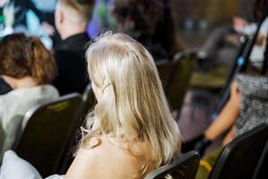Blond woman with layered hair sitting at event, facing crowd photo
