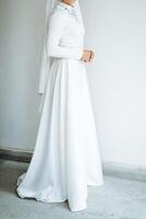 A woman, elegantly adorned in a stunning white dress and a delicate veil, stands gracefully in front of a pristine white wall, creating a beautiful and serene atmosphere around her photo