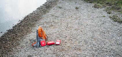 A backpack and a first aid kit can be seen laying on the ground next to the edge of a body of water, possibly indicating someone was preparing for an outdoor adventure nearby photo