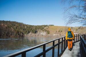 A solo solo trip with a backpack in a mountainous area, a guy walks on a bridge along the river, trekking in cool weather, a sunny day in nature. photo