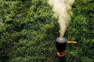 There is thick smoke billowing out from a pot placed in the middle of a vast open field, surrounded by lush green grass and various terrestrial plants, creating a striking visual event photo