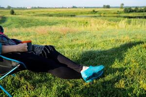 In a serene landscape, a woman is comfortably sitting in a chair with her feet propped up, surrounded by a vibrant field filled with lush grass and under a clear blue sky photo