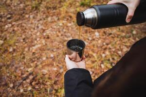 A person is in the act of pouring hot coffee from a thermos into a cup, showcasing relaxation and enjoyment in a serene setting. It is a delightful moment as they savor their drink photo