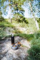In the peaceful forest, a kettle and two cups are placed on a boulder, amidst the beauty of nature, fostering a serene ambiance. The landscape enhances the tranquility of the setting photo