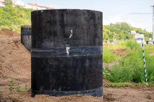 In the center of a wide dirt field, there is a large black cylinder prominently sitting, making it the focal point of the surroundings with grass and trees in the background photo