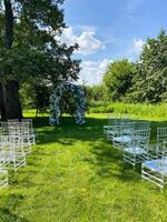 A row of white chairs is arranged in a grassy field for a wedding ceremony beneath the expansive sky. The setting includes elements like water, clouds, plants, and grass photo