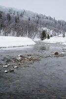 A serene river gracefully flows through a picturesque snowy forest, while majestic mountains stand tall in the background, together creating a breathtaking winter landscape that captivates photo