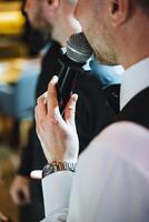A man in formal wear with a beard holding a microphone at an entertainment event photo