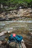 Backpack rests on rock by river, blending with grass and electric blue water photo