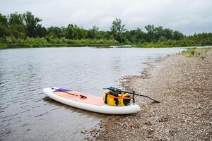 Inflatable Paddleboard with Yellow Bag and Paddle on Rocky Riverbank on Overcast Day, offering a tranquil setting for water sports among greenery and calm waters photo