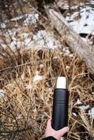 A person is seen holding a sleek black thermos in their hand while standing in the serene woods, surrounded by the beauty of nature and the tranquility of the environment photo
