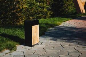 A beautifully crafted wooden trash can on a busy park sidewalk, surrounded by greenery, offers a convenient waste option while enhancing the parks aesthetics and natures beauty photo