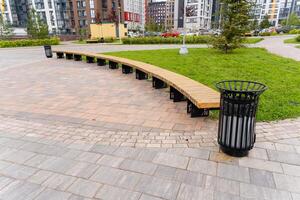 There is a wooden bench situated in a park right next to a trash can photo