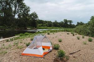 A tent is pitched on a sandy beach by a river, surrounded by natural landscape. The serene setting offers a peaceful escape with water, trees, grass, and a clear sky above photo