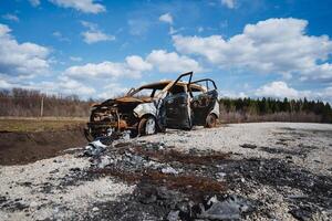 A burnt car is abandoned on the road in a field against the background of the sky, rusty iron, charred earth. photo