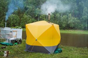 A vibrant yellow camping tent is set up by a lake in a serene forest, with smoke trailing from its chimney. Its an idyllic and peaceful scene of nature and exploration photo