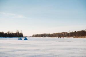 A diverse group of friends enjoys ice fishing together on a frozen lake, surrounded by the striking beauty of snow and shimmering ice underneath a chilly, clear blue sky overhead photo