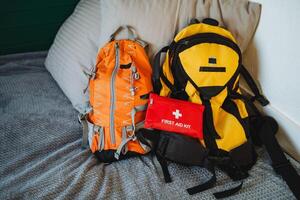 On a cozy bed, two backpacks wait, one vibrant yellow, alongside a neatly arranged first aid kit, ready for travel or outdoor adventures, ensuring accessibility for any situation photo