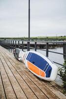 On a cloudy day, a paddleboard rests on a wooden dock by the waterfront, creating a serene coastal scene with marine elements. The setting exudes tranquility and beauty photo